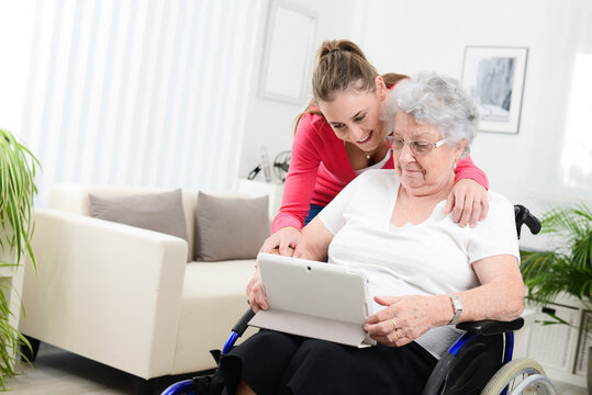 Cheerful young girl playing on internet with tablet computer and sharing time with old senior woman on wheelchair