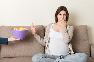 Pregnant woman sitting on the sofa refuses to eat unhealthy snacks. Stop to the junk food during pregnancy concept