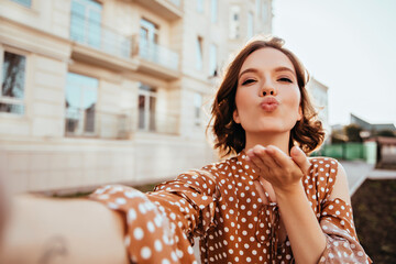 Blissful girl in vintage attire making selfie on the street. Gorgeous caucasian lady in brown outfit sending air kiss to camera. - Powered by Adobe
