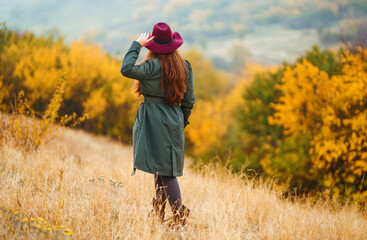 Elegant woman in coat with hat walks in autumn park. Stylish woman enjoying autumn weather in the meadow. Autumn fashion. Rest, relaxation, lifestyle concept.
