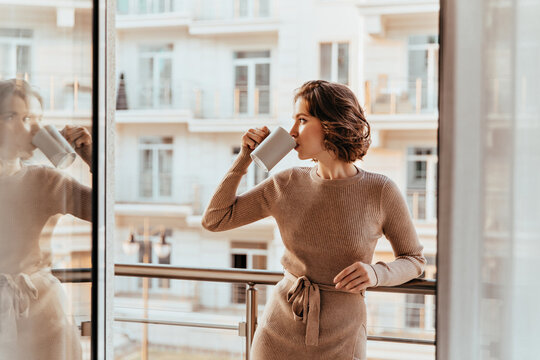 Joyful young woman drinking coffee and looking at city. Indoor photo of pleased curly girl in brown dress spending morning at balcony.