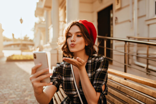 Glamorous brunette woman sending air kiss while making selfie in sunny day. Graceful french female model holding smartphone and taking picture of herself.
