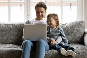 Fototapeta premium Happy mommy cuddling small adorable kid son, using funny children educational applications on computer. Smiling affectionate mum enjoying watching funny cartoons with small child boy at home.