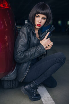 Beautiful brunette sexy spy agent (killer or police) woman in leather jacket and jeans with a gun in her hand running after someone, to catch him on parking