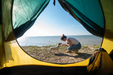 View from a tourist tent on a woman playing with a dog on the seashore. The girl set up camp in the countryside.