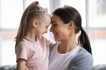 Head shot happy little blonde girl wearing princess crown, touching foreheads with caring young beautiful mother, enjoying tender sweet family moment together indoors, daycare pastime concept.