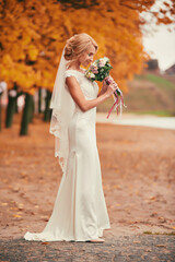 Fototapeta na wymiar Beautiful bride in white long dress is smelling wedding bouquet in autumn park on wedding day