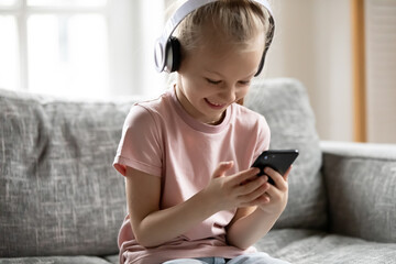 Overjoyed little adorable blonde girl wearing wireless headphones, watching funny video in social...