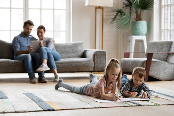 Happy little friendly children siblings lying on floor carpet involved in drawing pictures in paper album while smiling young parents relaxing on sofa using digital computer tablet in living room..