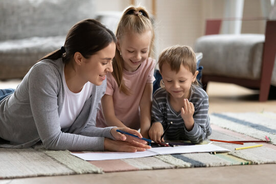 Affectionate loving young mother enjoying painting pictures in paper album with adorable small children siblings. Smiling babysitter teaching kids drawing, lying together on comfortable floor carpet.