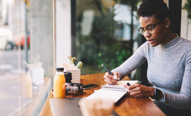 Pensive talented attractive afro american female journalist in glasses thinking over creative solutions for making article working in cafe, charming skilled designer noting ideas for start up