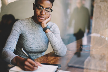 Attractive afro american experienced owner of business startup in glasses and casual wear preparing report. Concentrated student writing checklist in notebook sitting in modern interior coffee shop