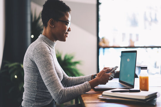 Smiling afro american female copywriter satisfied with working process in cafe interior synchronizing files with smartphone using wifi connection, attractive freelancer receiving job offer on email