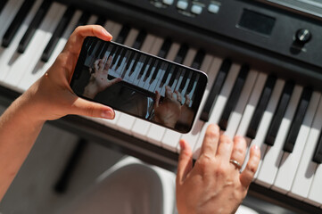 A girl watches an online music lesson on a smartphone and learns to play the piano. Close-up of woman's hands on keys.