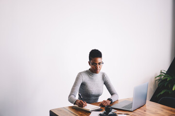 Concentrated Afro-American female designer creating sketch in notebook concentrated on ideas, professional businesswoman in eyewear planning projects making notes sitting on promotional background