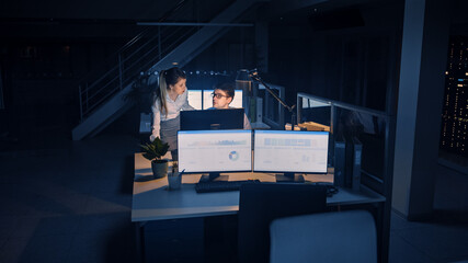 Confident Businessman Uses Desktop Computer, His Female Project Manager Explains Specific Tasks, Account Handling and Strategic Moves. Professional People Late at Night in Big Corporate Office