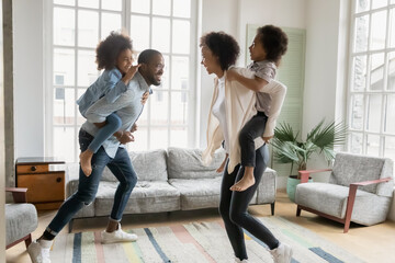 Overjoyed little ethnic children piggyback playful young mother and father play together in living room, excited small african American kids enjoy funny ride game with playful ethnic parents at home