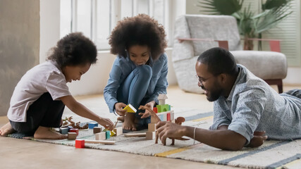 Loving young african American father sit on floor in living room play with toys blocks with small...