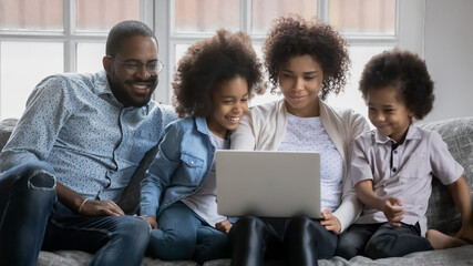 Happy african American family with two little children sit relax on couch in living room using laptop together, smiling biracial young parents with small kids watch video on computer at home