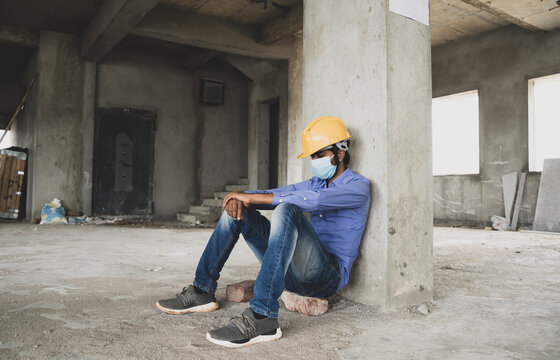 Construction Worker Sitting Sad And Lonely At Job Site Wearing A Medical Mask With Hardhat To Prevent Covid-19 Spreading, Concept Of Unemployment, Economic Crisis Job Loss During Coronavirus Crisis.