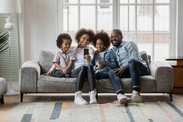 Smiling young african American family with two small children sit on couch make self-portrait picture on cellphone, happy biracial parents with little kids have fun pose for selfie on smartphone