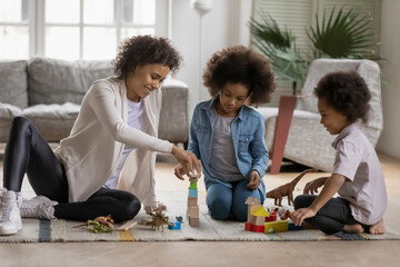 Fototapeta premium Playful young african American mom or nanny sit on floor at home play with building bricks with two little children, happy biracial mother have fun construct with blocks with small kids in living room