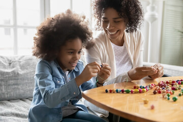 Smiling young african American mom or nanny sit at table play make bracelets with little girl...