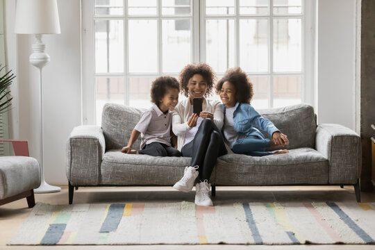 Overjoyed young biracial mother sit relax on couch at home with little children make self-portarit picture on cell, smiling african American mom and two small kids pose for selfie on smartphone