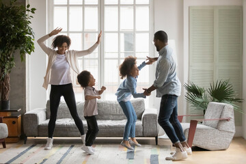 Overjoyed african American family with two little kids have fun dancing jumping together in living room, happy young ethnic parents with small children engaged in funny activity at home on weekend