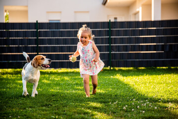 Baby girl running with beagle dog in garden on summer day. Domestic animal with children concept.