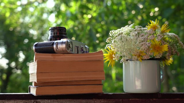 Closeup view 4k still life video footage of old vintage photo camera laying on stack of old paper books on brown wooden background. Beautiful valley flowers arranged in blue enamel mug.