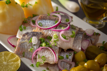 Pickled herring slices with red onion, fermented cucumber, potatoes, olives and lemon in a white plate on a dark wooden table. Close-up
