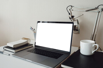 Modern stylish personal laptop with a blank white screen. Freelance artist or designer workplace. Clean display. Table lamp. Paint brushes. Notebooks for drawing and writing. White cup.