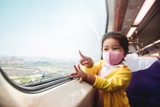 Travel in New Normal Lifestyle Concept. Happy Kids wearing  Surgical Protection Mask inside a Train while Traveling with her Parent. Sitting by Wide Glass Window to take a View