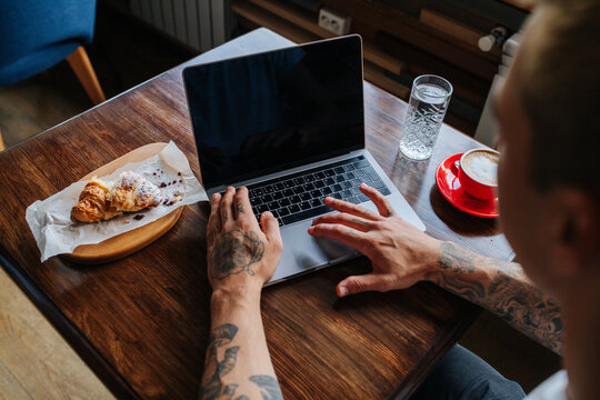 Man Working On His Laptop Behind The Table In A Cafe. Over A Shoulder View.