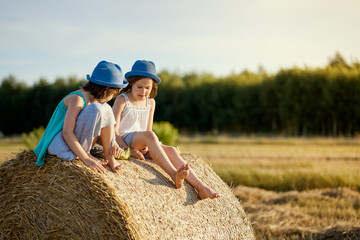 Naklejka na ściany i meble two charming girls are sitting on a roll of mown rye in a field