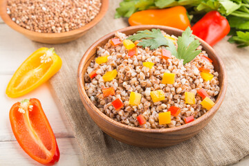 Buckwheat porridge with vegetables in wooden bowl on a white wooden background and linen textile. Side view, close up, selective focus.