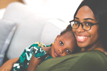 Happy young African American mother having fun with daughter child girl playing and hugging in bedroom at home