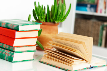 Cozy home interior decor: stack of books, plants in pots, open book on a white table. Distance home education.Quarantine concept of stay home.