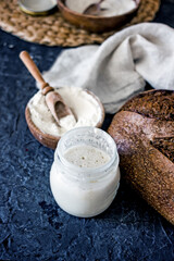 Bread starter sourdough in a glass jar on a dark background, near bread and flour, vertical background, close-up