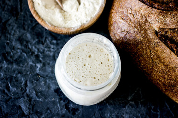 Bread starter sourdough in a glass jar on a dark background, near bread and flour, vertical background, close-up
