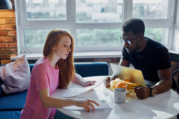 african man work on laptop, his wife checking utility bills and house maintenance documents, they...