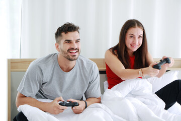Smiling young Caucasian lover Couple hand holding joysticks and playing game play station together in bedroom. Lifestyle and relationship of married life on holiday Concept.