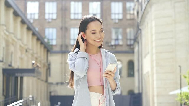 Young smiling woman standing with smartphone at the street and listening to music.