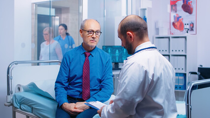 Senior man consulting with doctor sitting on hospital bed. Ill aged patient seeking medical advice for disease prevention from general practitioner in modern private clinic, doctor diagnosis