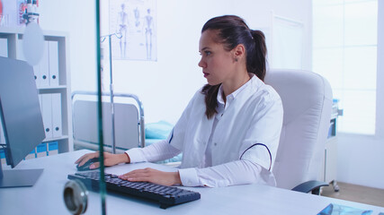 Medic wearing white coat in hospital cabinet working on computer. View through glass wall. Assistant giving patient radiography. Medic examining patient radiography before giving a treatment to cure