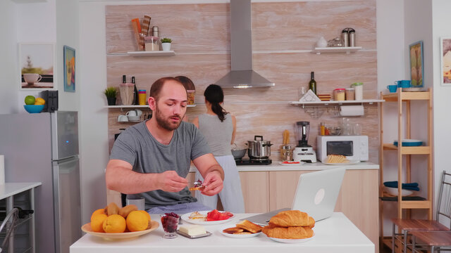 Entrepreneur working on laptop in kitchen and drinking coffee while wife is cooking breakfast. Freelancer work online via internet using modern digital technology