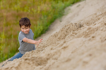 A happy boy plays on a pile of sand against a background of green grass in summer. The concept of distancing oneself from people because of an epidemic. Activity outdoors.