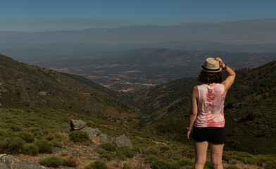 Naklejka premium Attractive young female standing on the top of a rock witnessing the landscape of the north of Extremadura, Spain, while wearing a reflex camera to take photos
