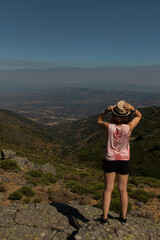 Naklejka premium Attractive young female standing on the top of a rock witnessing the landscape of the north of Extremadura, Spain, while wearing a reflex camera to take photos